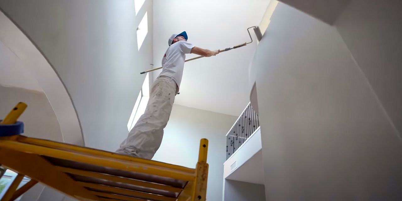 Painter on a ladder rolling paint onto a tall stairwell wall