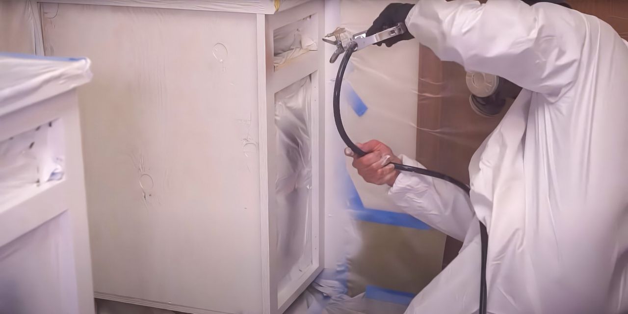 Painter in a protective suit spraying a kitchen cabinet