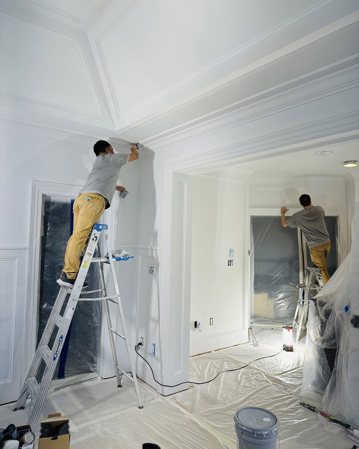 Two painters on ladders painting a room with the floor covered in plastic